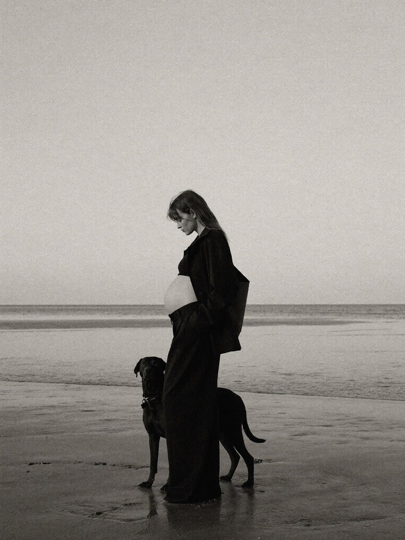 Black-and-white editorial image of a pregnant woman in a long coat on a beach.