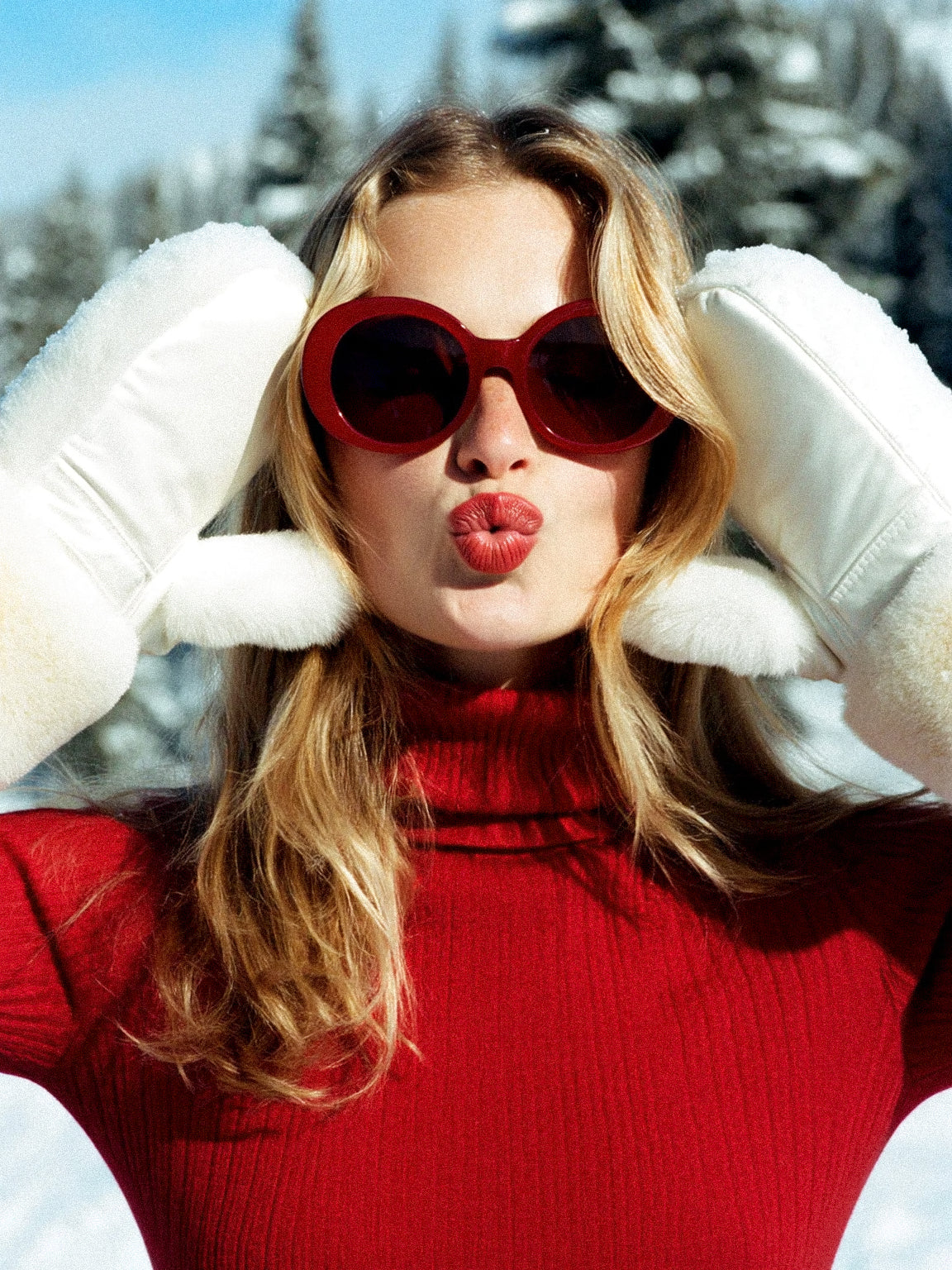 A woman in a red turtleneck and round red sunglasses, white mittens at her ears, in snow.