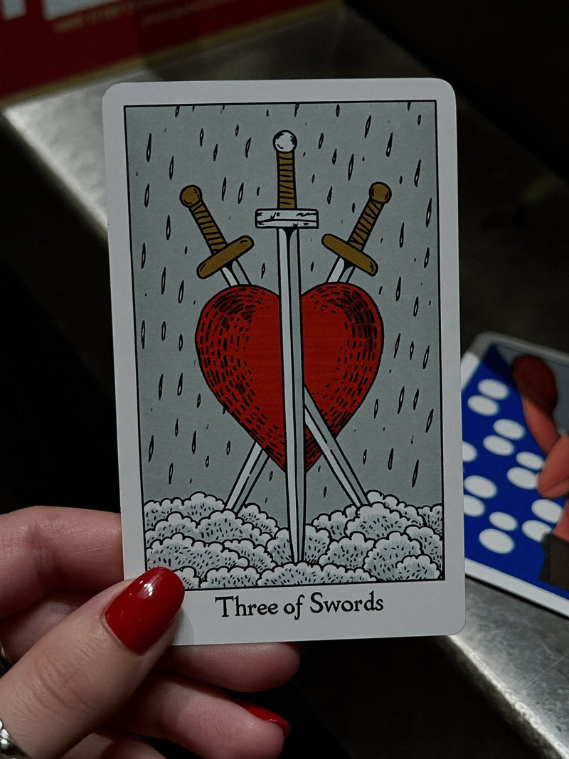 A hand holding a Three of Swords tarot card, a red heart pierced by three swords against a grey sky.