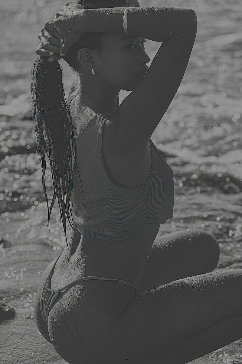 Black-and-white editorial photo of a woman kneeling in shallow surf, arms raised to her hair.