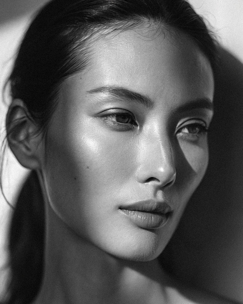 Editorial portrait of a woman with smooth, even-toned skin in dramatic side-lighting. Black-and-white.