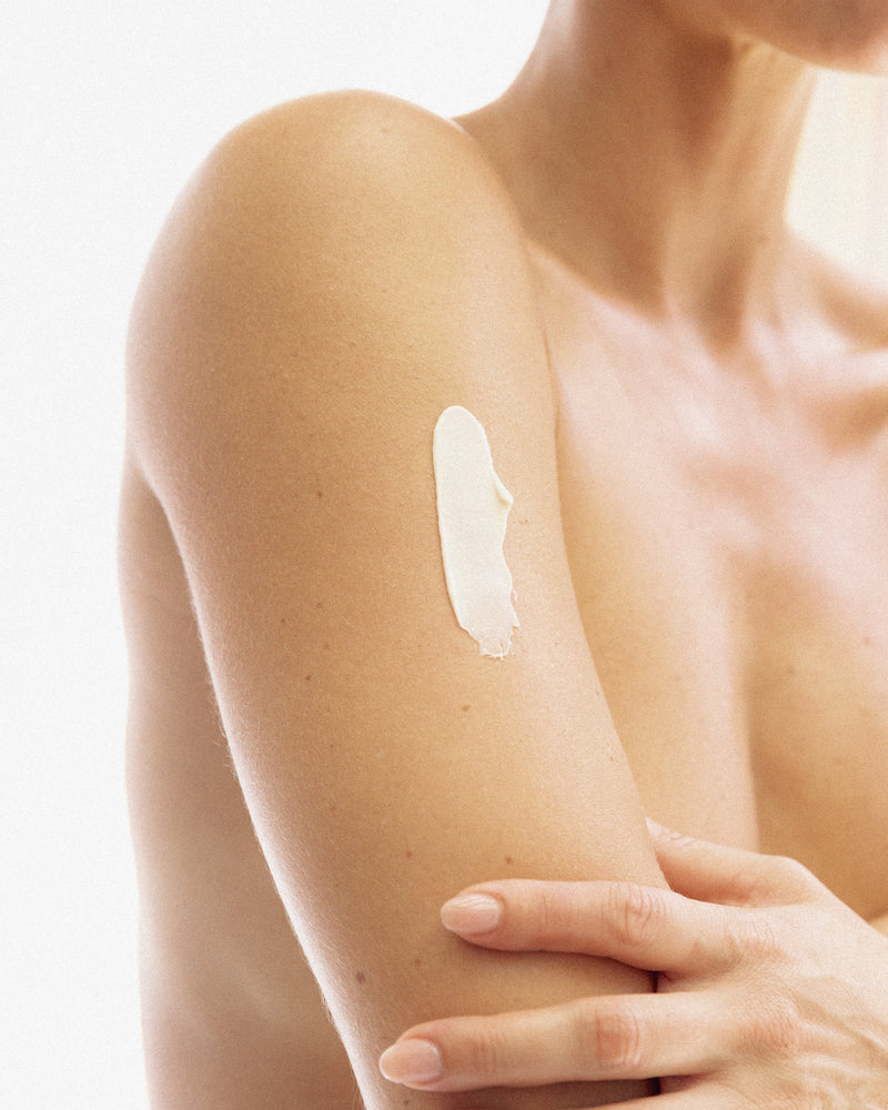 Woman's bare shoulder and upper arm with a dollop of white skincare cream on the upper arm in soft daylight.