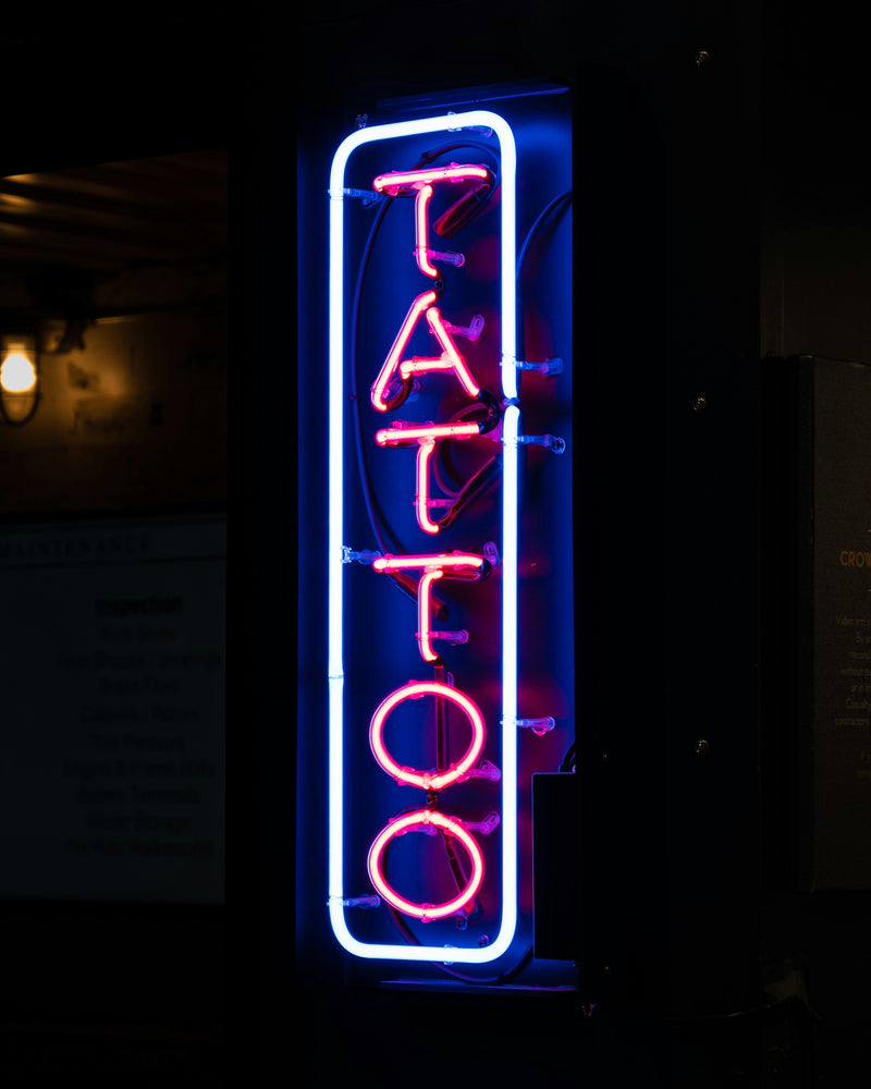 A glowing neon 'Tattoo' sign. The cost difference between applying a tattoo and removing one can be significant.