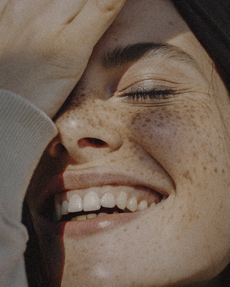 Young woman with freckles, smiling with eyes closed in warm sunlight.