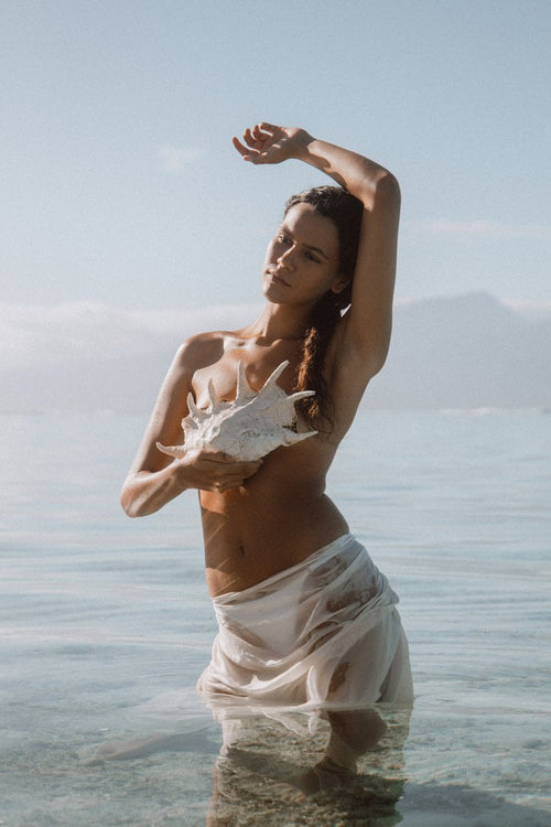 Woman standing in shallow water holding a large white shell to her chest, one arm raised, sunlit skin against a pale sky.