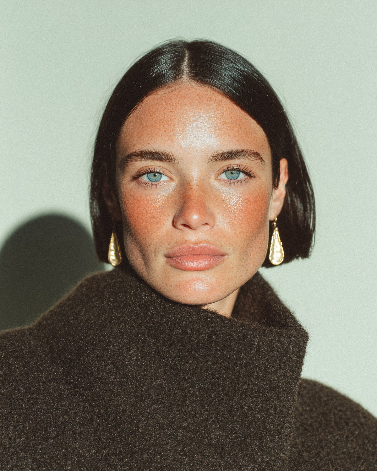 Editorial portrait of a young woman with freckled skin, dark bobbed hair, and gold drop earrings, against a sage green background.