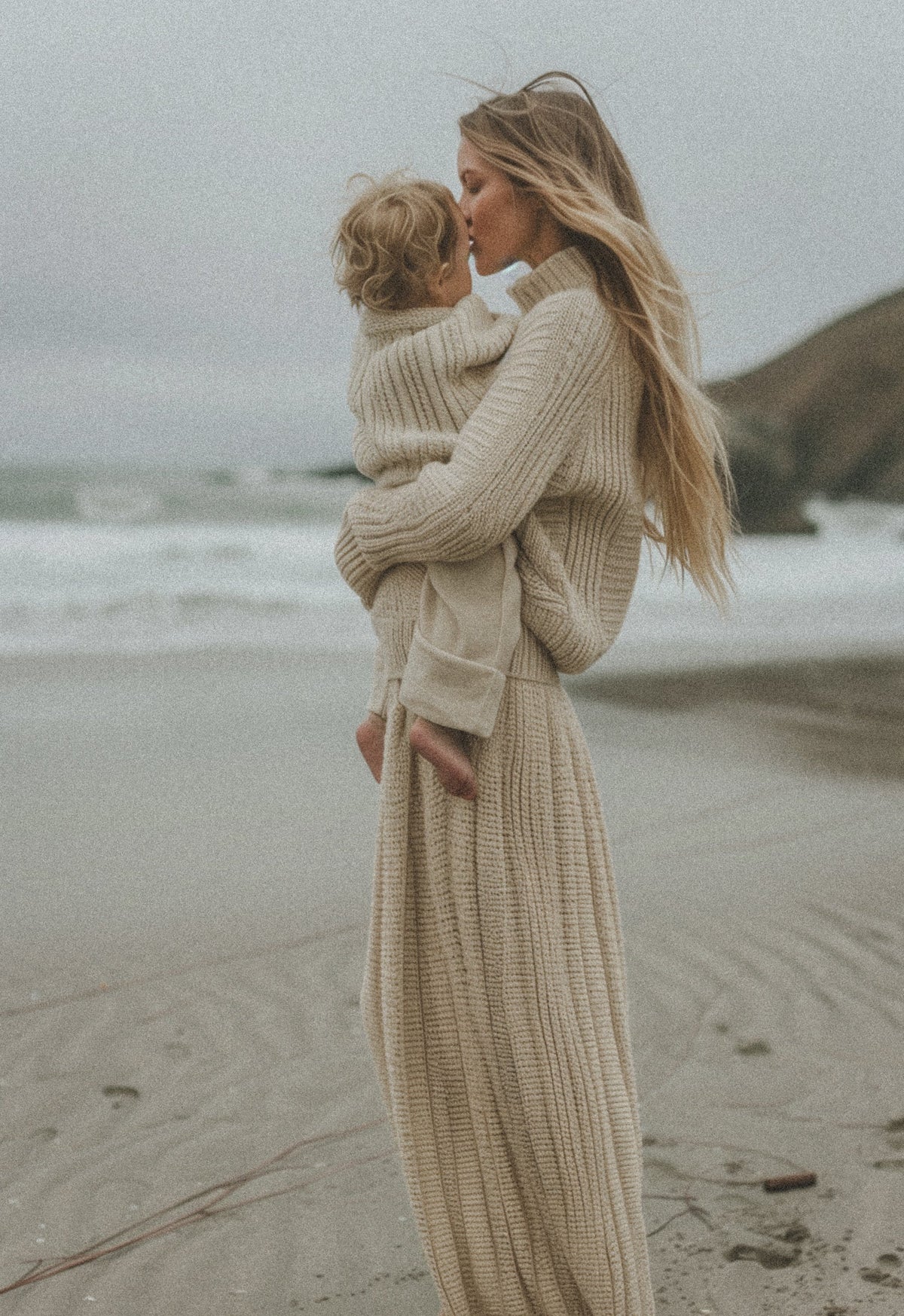 Mother in a cream knit cradling her baby on a beach, evoking postpartum pelvic-floor recovery.