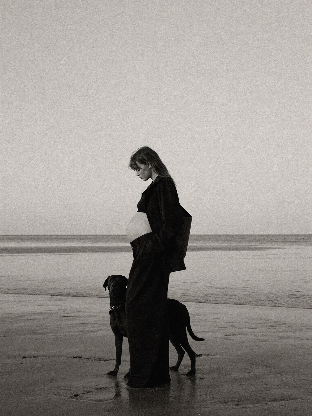 Black-and-white editorial image of a pregnant woman in a long coat on a beach.