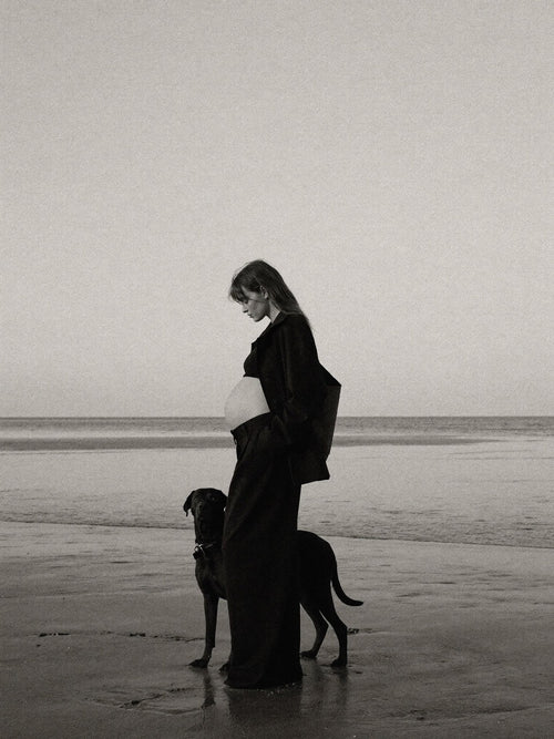 Black-and-white editorial image of a pregnant woman in a long coat on a beach.