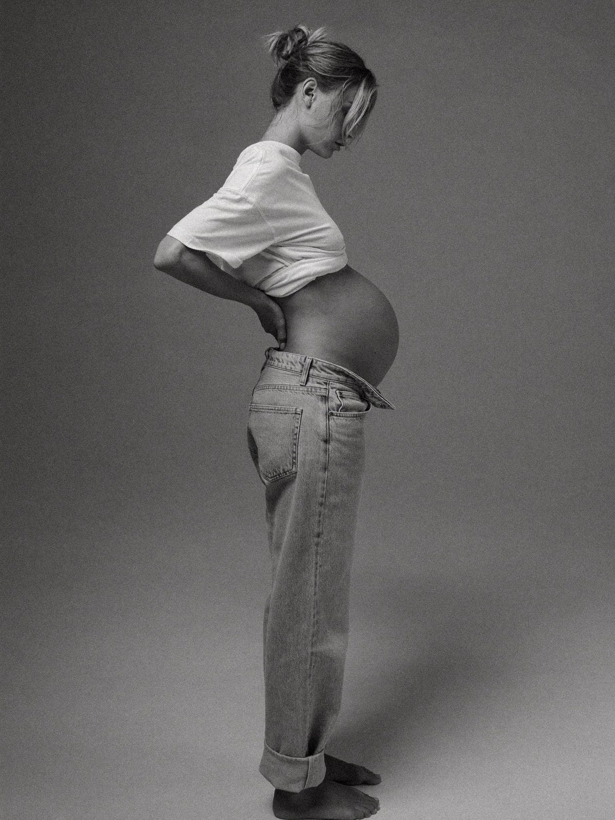Pregnant woman in jeans, side view, with a hand supporting her lower back.