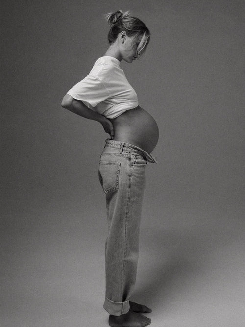 Pregnant woman in jeans, side view, with a hand supporting her lower back.