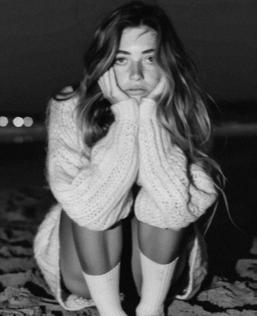 A young woman in a knit jumper, hands to her face, contemplative, in black and white.