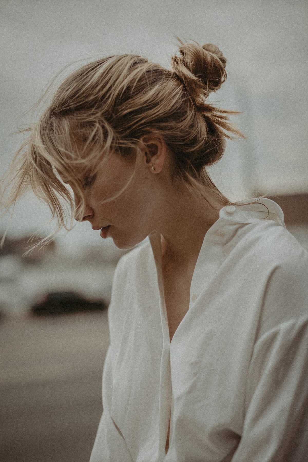 A woman in a white shirt, hair in a loose bun, looking down, soft and contemplative.