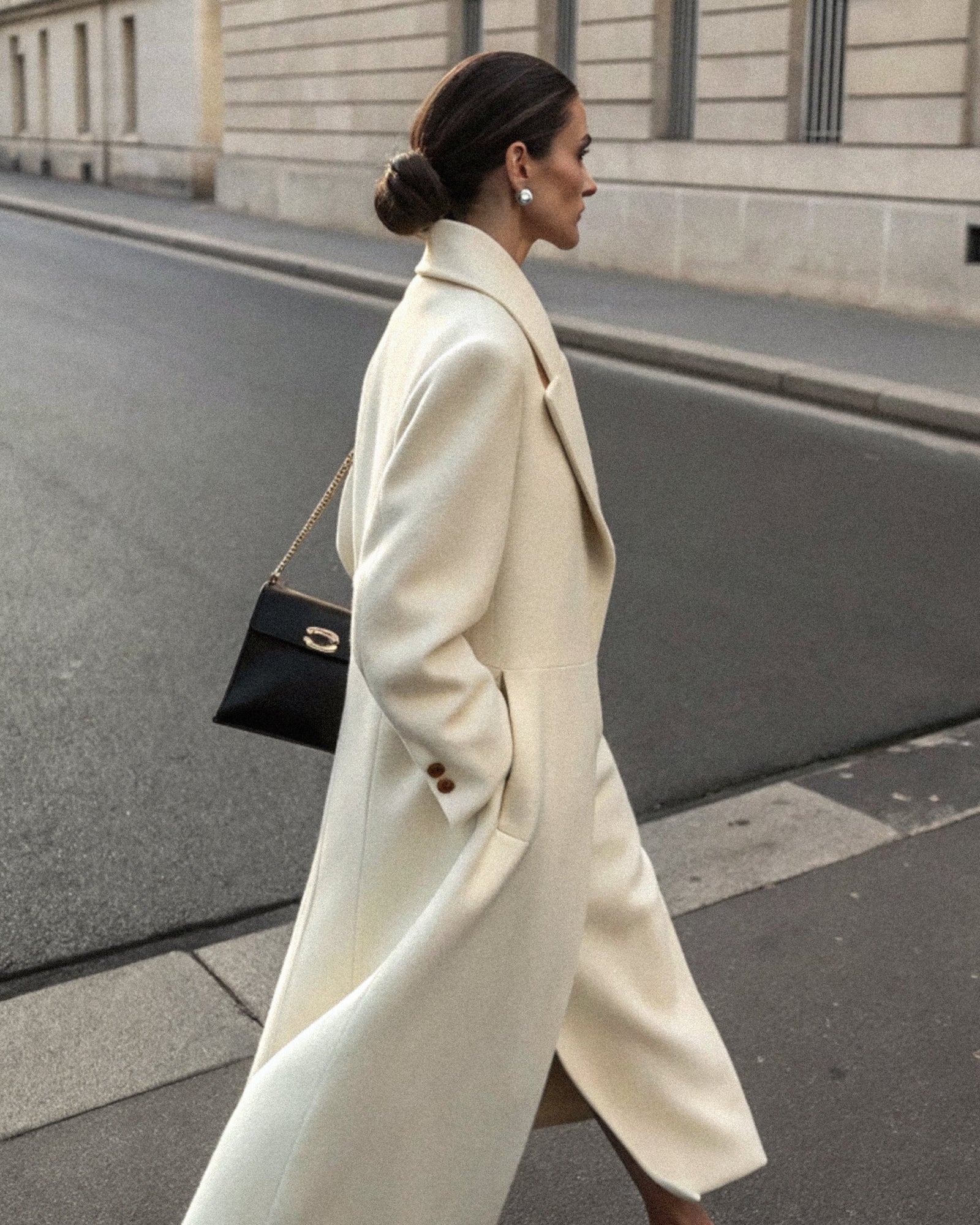 An elegant woman in a cream coat walking a quiet street, seen in profile.