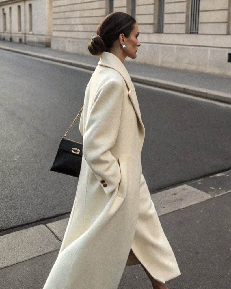 An elegant woman in a cream coat walking a quiet street, seen in profile.