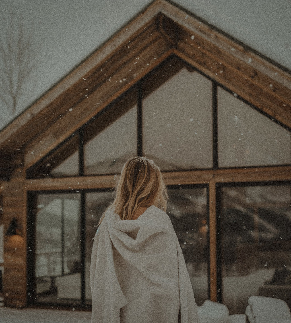 A woman wrapped in a blanket facing a cabin with large windows, snow falling.