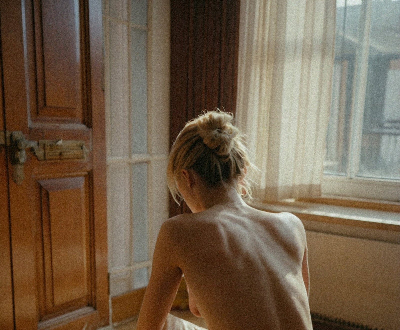A woman seen from behind, bare shoulders and hair up, by a window in warm light.