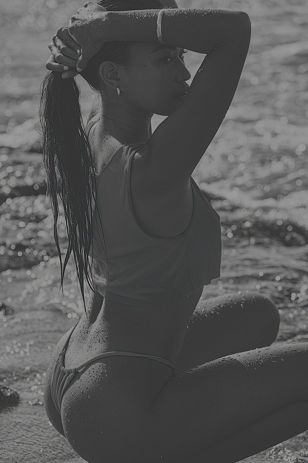 Black-and-white editorial photo of a woman kneeling in shallow surf, arms raised to her hair.