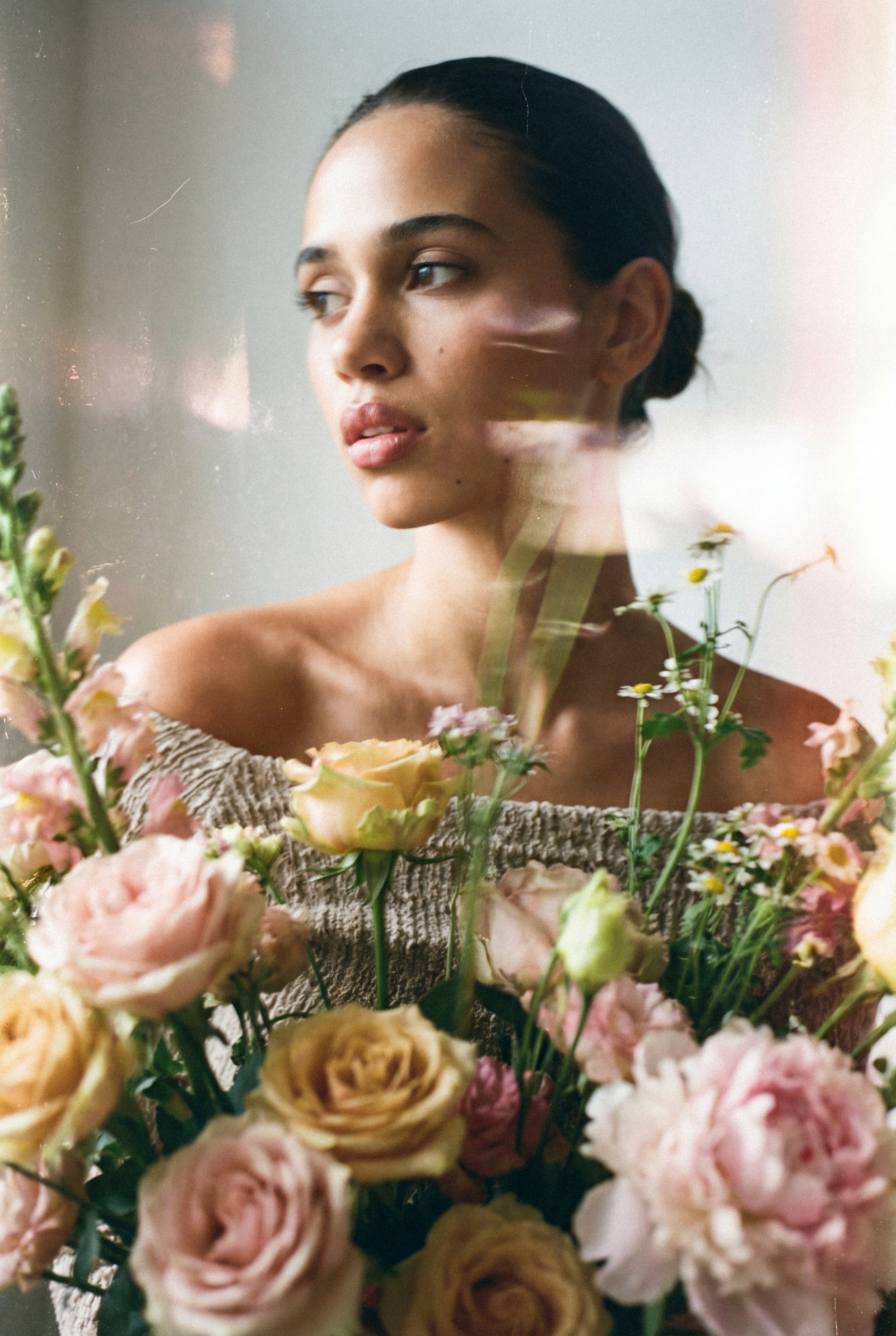 Editorial portrait of a woman with luminous, glowing skin in soft natural light, behind an arrangement of pink and cream roses.